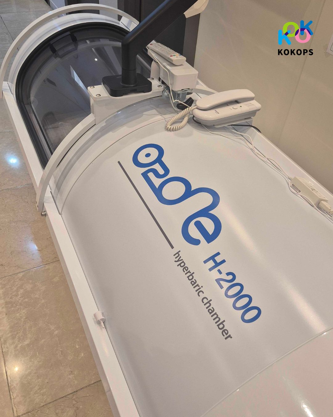 高圧酸素治療装置「O2 One H-2000」の本体上部のアップ写真。装置の上にはモニター用のアームや、外部と連絡をとるための白い電話機（インターホン）が設置されている。