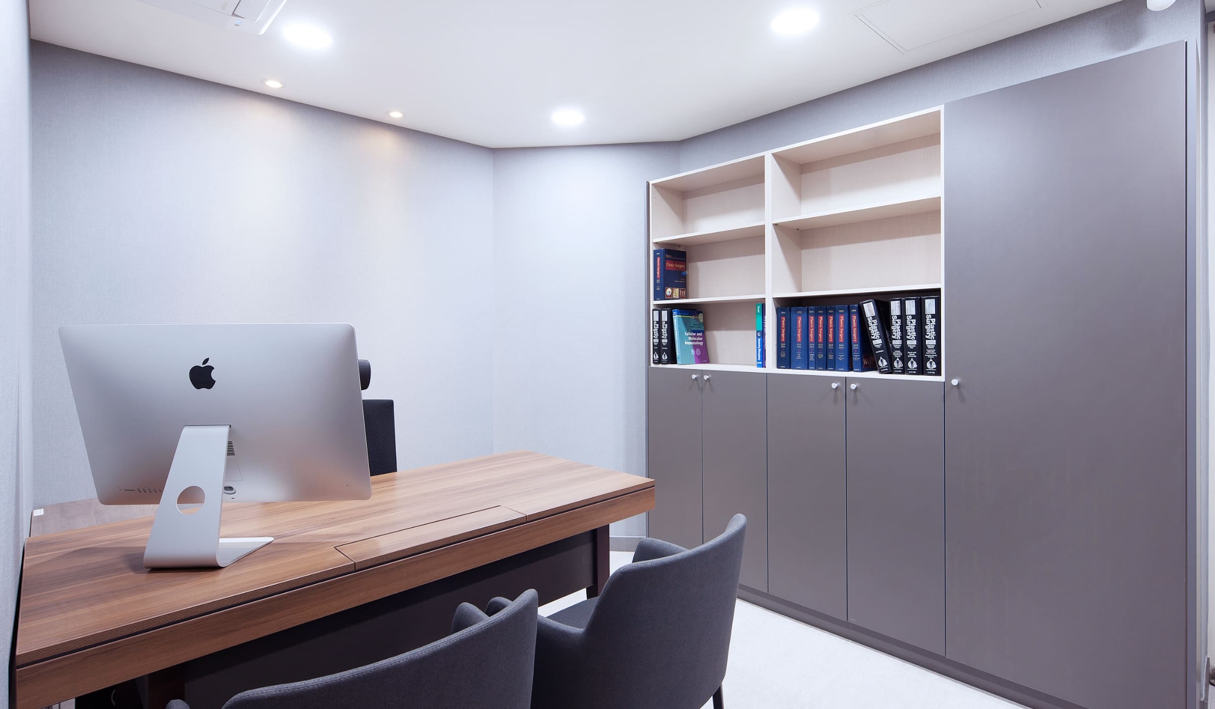 Consultation room in a plastic surgery clinic with a wooden table, gray chairs, and a bookshelf.